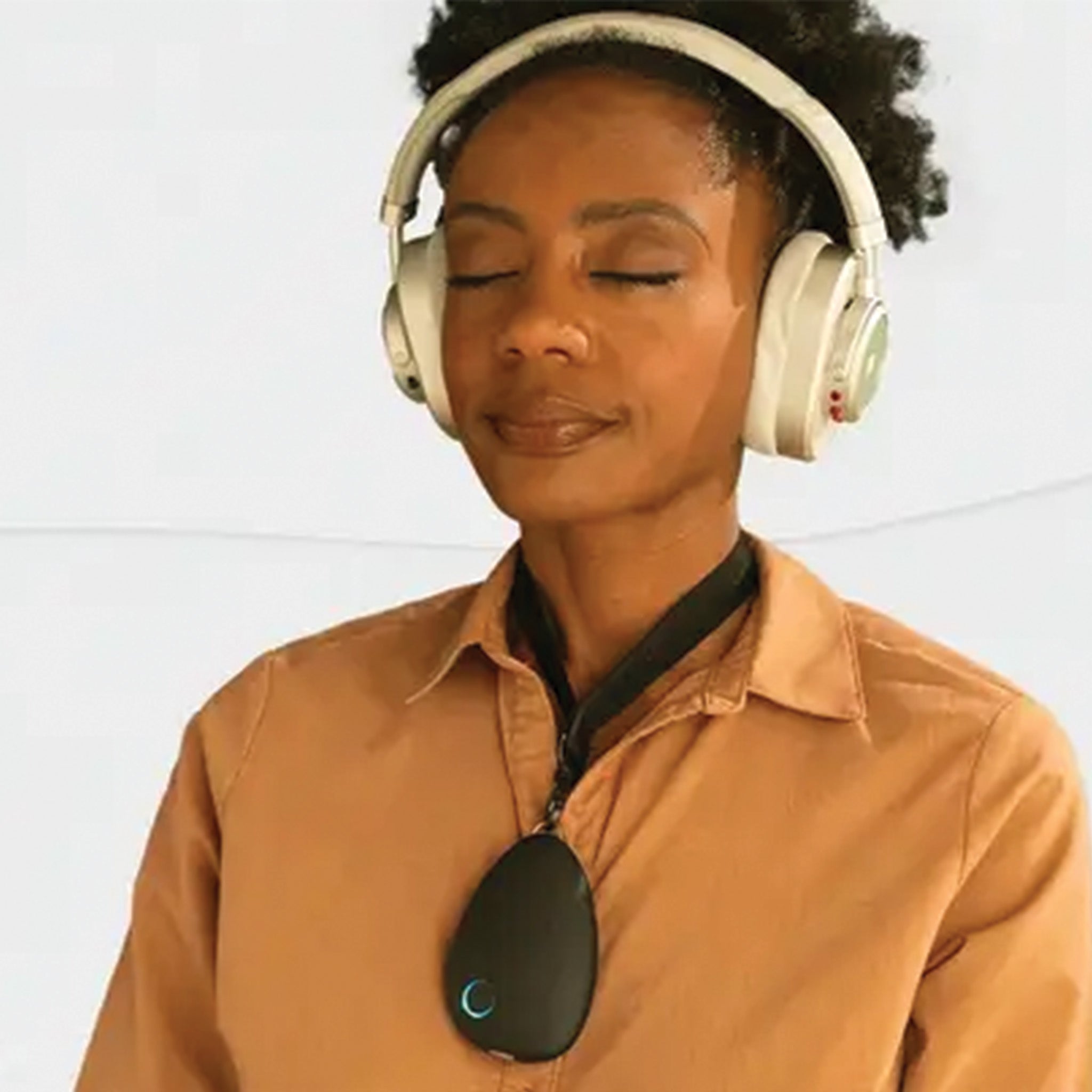 A lady with black skin with a sensate on her chest like a pendant. Her eyes are closed and she has cream headphone on.using a sensate